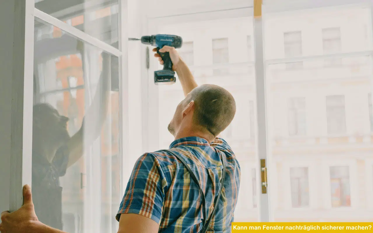 Ein Handwerker in einem blau-orange karierten Hemd steht vor einem großen weißen Fenster und bohrt mit einem Akkubohrschrauber in den oberen Teil des Rahmens, um Sicherheitstechnik zu montieren.