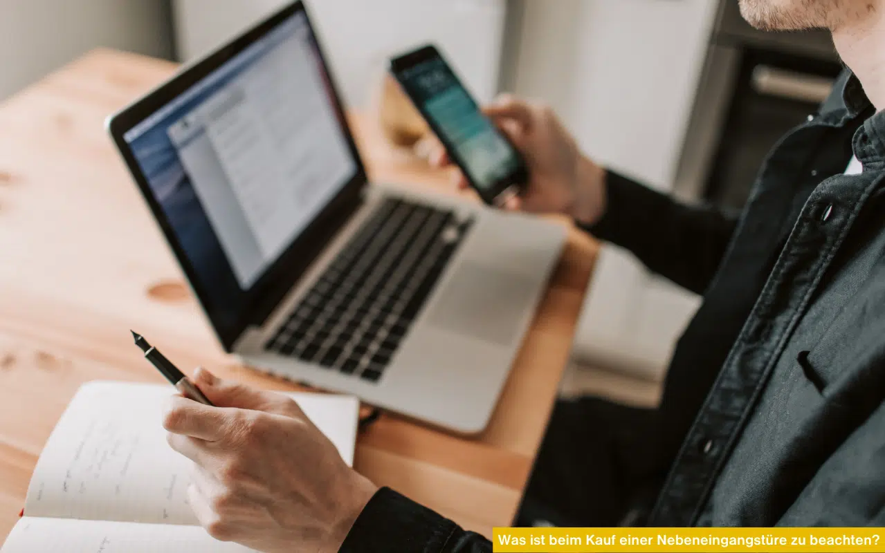 Ein Mann am Schreibtisch nutzt Laptop und Smartphone für eine Recherche; vor ihm liegt ein offener Notizblock.