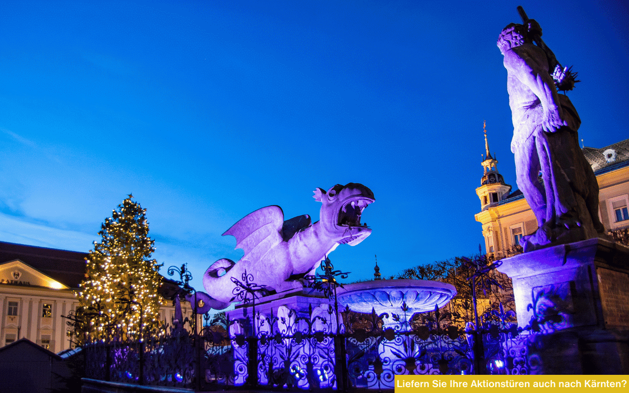 Der beleuchtete Lindwurmbrunnen in Klagenfurt bei Nacht mit Weihnachtsbaum im Hintergrund.