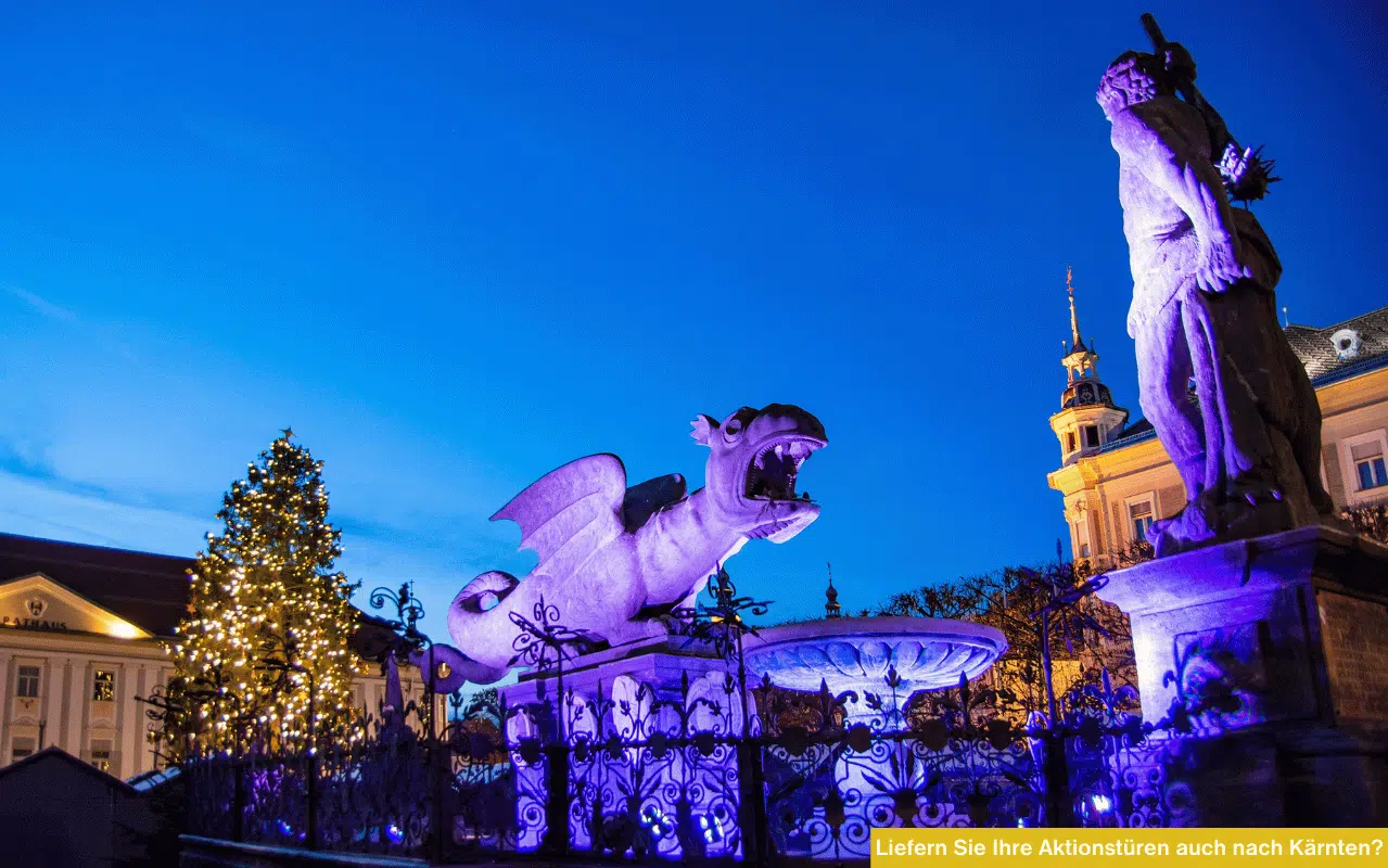 Der beleuchtete Lindwurmbrunnen in Klagenfurt bei Nacht mit Weihnachtsbaum im Hintergrund.