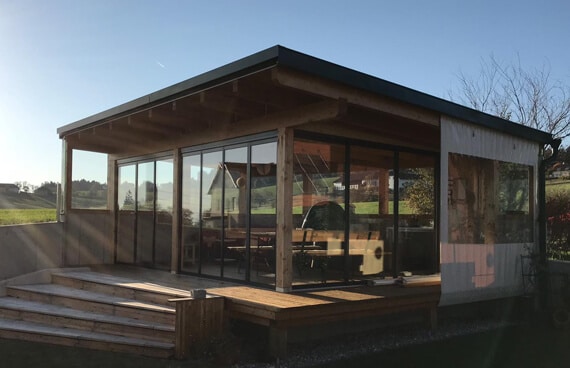 Modernes Gartenhaus mit Pultdach und großer Glasfront, davor Holzstufen und Terrasse, im Hintergrund Wiese und Häuser.