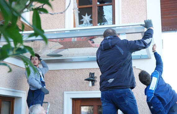 Das Bild zeigt mehrere Monteure, die eine Glasüberdachung an einer Hausfassade montieren. Die Glasplatte wird gemeinsam angehoben und ausgerichtet – typische Montagesituation im Außenbereich.