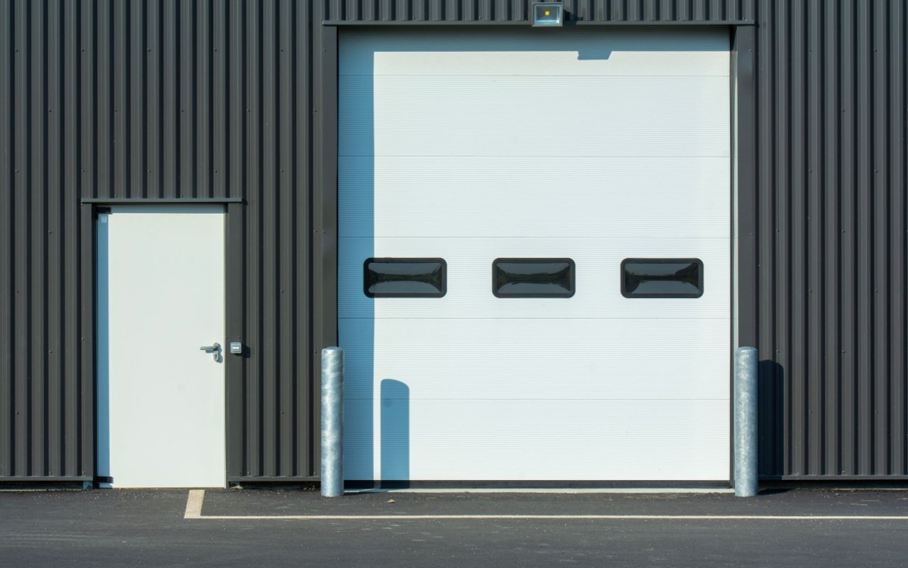 Weiße Nebeneingangstür neben einem weißen Sektionaltor in einer dunklen Blechfassade, davor zwei Rammschutzpfosten
