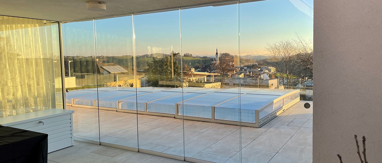 Rahmenlose Sunflex-Glasschiebewand mit fünf Glasflügeln auf einer Terrasse bei Sonnenuntergang - offene Glasfront mit Blick auf Landschaft.