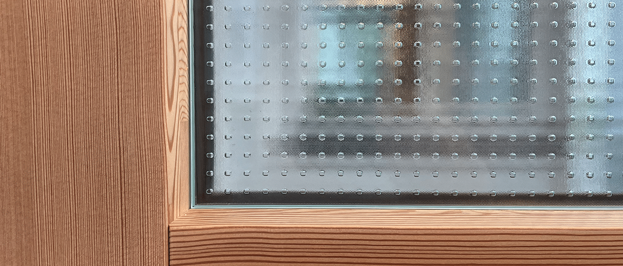 Das Bild zeigt ein Holz-Alu-Fenster in Lärchenholz mit Master-Carré Glas. Das karierte Muster des Glases erzeugt interessante Reflexionen und bietet gleichzeitig Sichtschutz in einem hellen Raum.