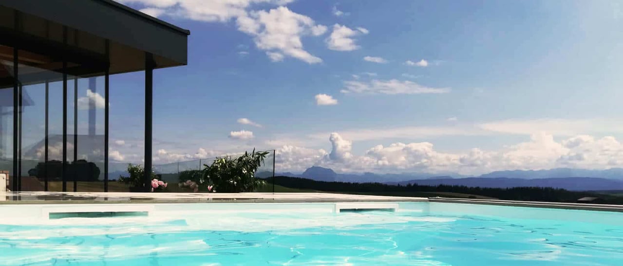 Das Bild zeigt ein modernes Poolhaus mit klarer Glasarchitektur und Infinity-Pool in traumhafter Landschaft. Es vermittelt pures Sommerfeeling, Ruhe und Luxus - der perfekte Ort zum Entspannen und Genießen.