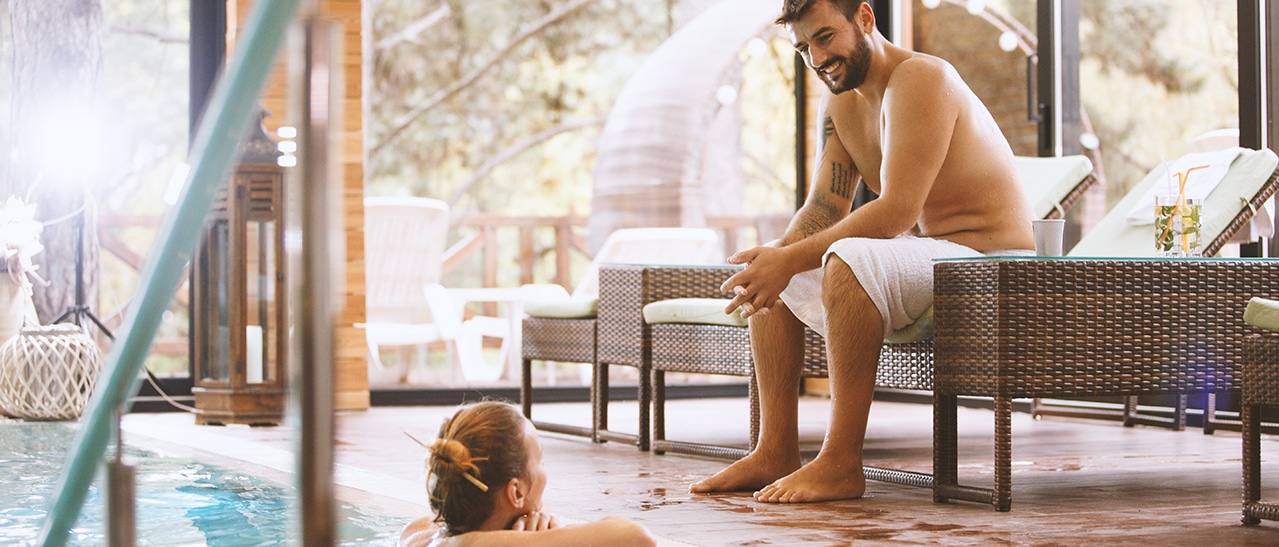 Eine stilvolle Poolüberdachung aus Glas verwandelt den Wintergarten in eine Wellnessoase. Ein Mann sitzt am Beckenrand, während eine Frau im Wasser schwimmt. Das warme Licht schafft eine ruhige, behagliche Stimmung.