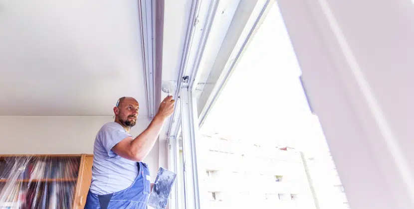 Ein Fachmonteur von Fenster-Schmidinger in blauer Arbeitskleidung montiert präzise ein neues weißes Fenstersystem in einem hellen Wohnraum.