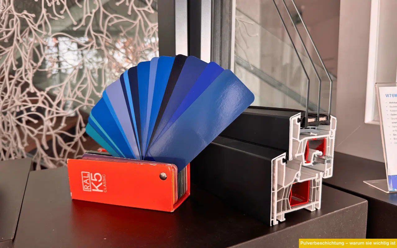 Das Bild zeigt ein aufgeschlagenes RAL-Farbfächerbuch mit verschiedenen Blautönen, das an einem Kunststoff-Aluminiumfensterprofil mit dunkler Pulverbeschichtung lehnt.