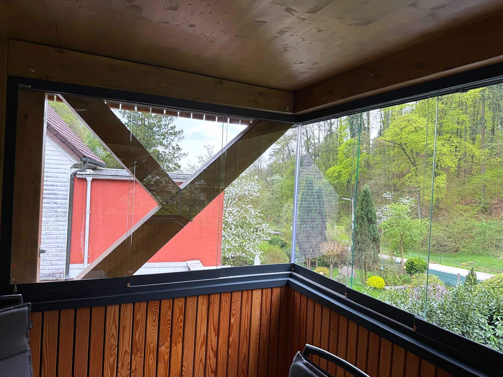 Rahmenlose Ganzglas-Schiebefenster an der Holzkonstruktion eines Balkons, Ecke ohne Steher, Diagonalstrebe außen, Blick über den Garten, Oberösterreich