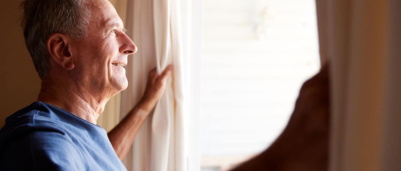 Älterer Mann lüftet früh Fenster für Kühlung. Tipps für kühle Wohnung im Sommer trotz Hitze.