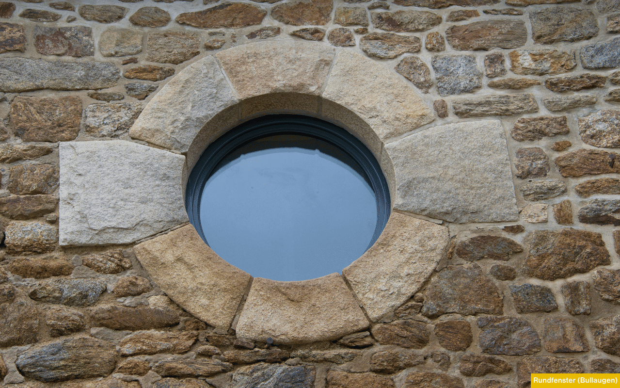 Sonderformen und Architekturfenster als ein rundes Fenster mit grauem Rahmen was in eine Natursteinmauer montiert ist und wirkt wie ein Bullauge.