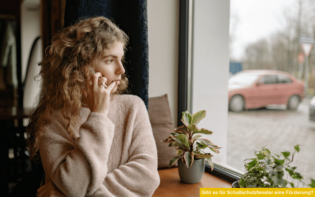 Frau telefoniert ungestört am Fenster, während draußen ein Auto vorbeifährt - Hinweis auf Förderung für Schallschutzfenster.