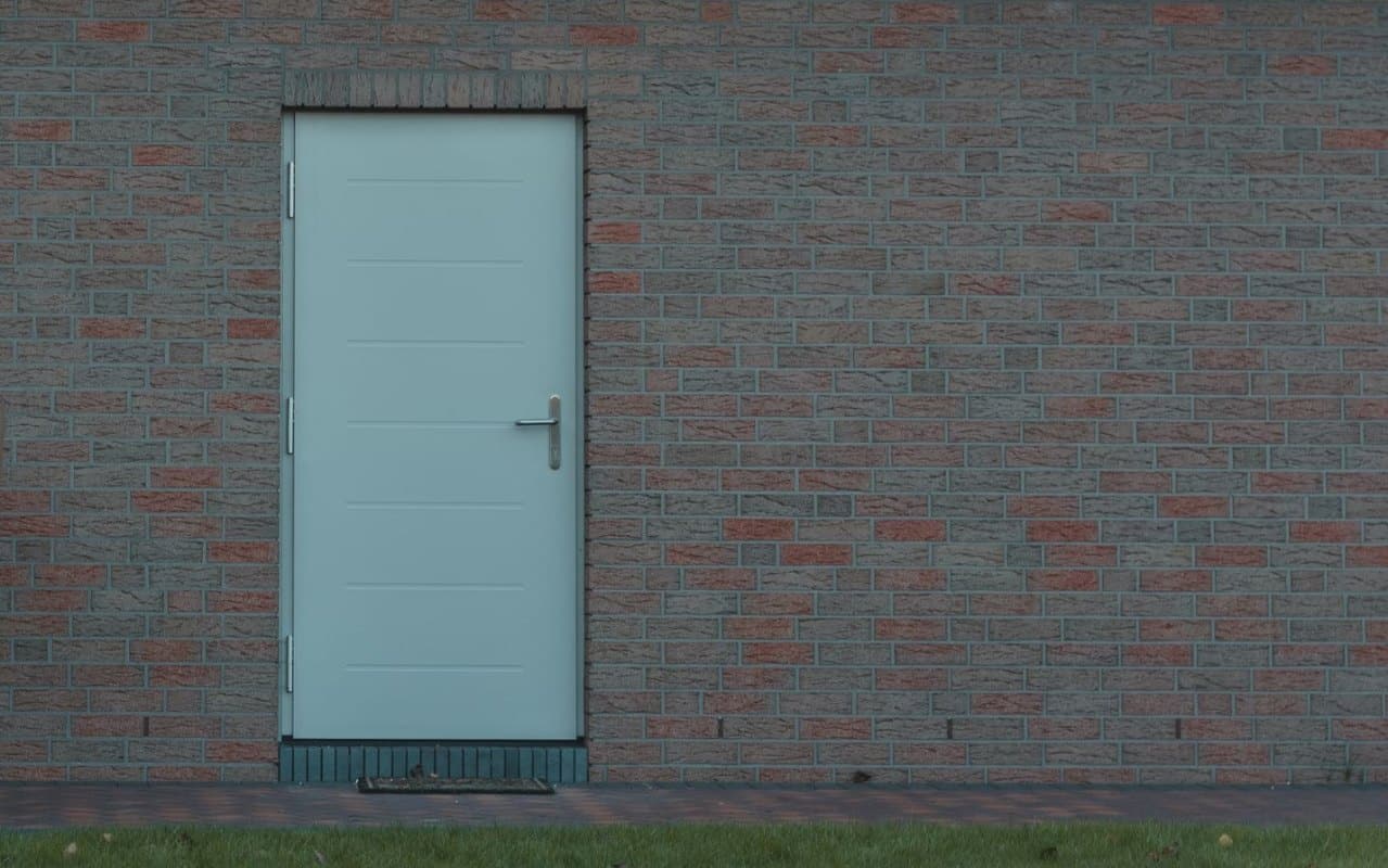 Schlichte hellgraue Nebeneingangstür mit silbernem Drücker in einer Ziegelwand, davor Rasen