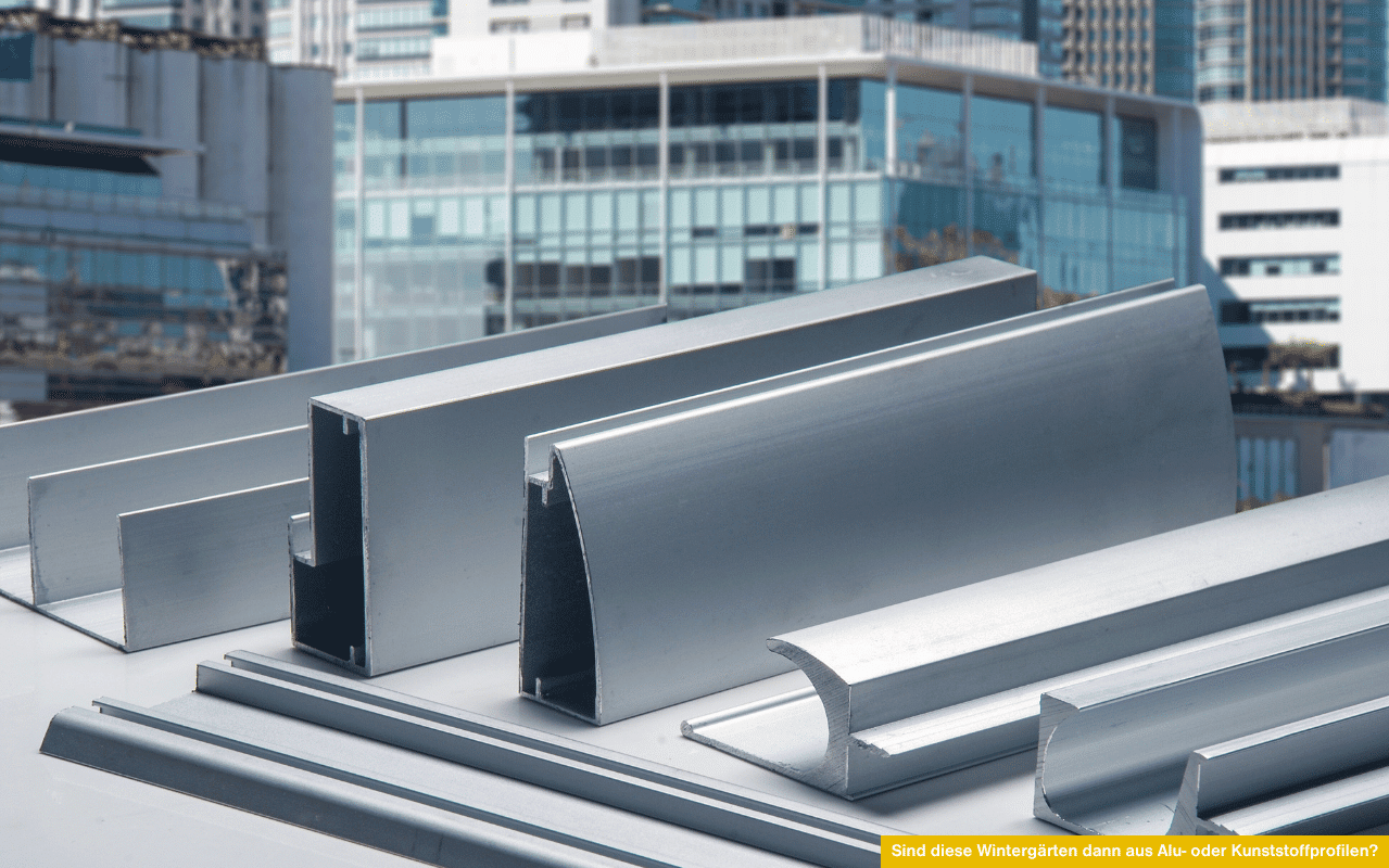 Das Bild zeigt verschiedene Aluminiumprofile in unterschiedlichen Formen und Größen auf einer hellen Fläche. Im Hintergrund sind moderne Gebäude zu erkennen. Das Foto symbolisiert die stabile Grundkonstruktion von Wintergärten aus Aluminium.