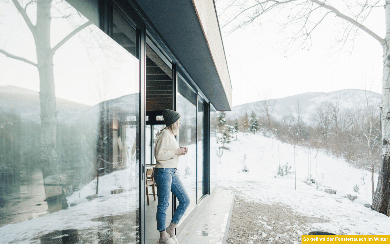 So gelingt der Fenstertausch im Winter Tipps zum Fenster im Winter tauschen Frau steht vor moderner Glasfront im verschneiten Garten.