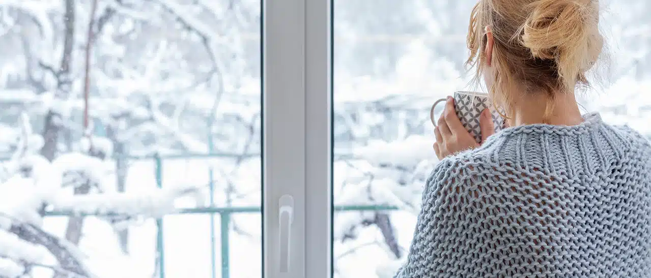 Frau wechselt Fenster im Winter und genießt die verschneite Landschaft – Tipps zum Fenster im Winter tauschen und ob ein Fenstertausch bei Minusgraden überhaupt möglich ist.