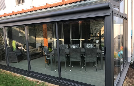 Das Bild zeigt die Außenansicht eines modernen Sommergartens, der an ein Wohnhaus in Österreich angebaut wurde. Die Konstruktion besteht aus anthrazitfarbenen Aluminiumprofilen und großflächigen Glasschiebeelementen.