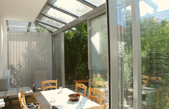 Ein moderner Sommergarten ist als heller Essbereich eingerichtet. In der Mitte steht ein Holztisch mit passenden Holzstühlen. Durch die großen Glasfronten fällt viel Licht ein und gibt den Blick auf den grünen Garten frei.