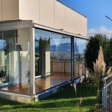 Sommergarten Erfahrungen und Anbieter: Verglaster Sommergarten mit Holzfußboden in sonniger Hanglage. Im Hintergrund idyllischer Bergblick und ein Wohngebiet mit Einfamilienhäusern.