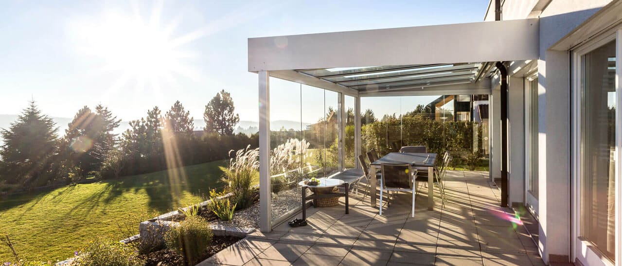 Moderner Sommergarten mit weißer Aluminiumkonstruktion, Glasdach und Glasschiebewänden auf einer sonnigen Terrasse.