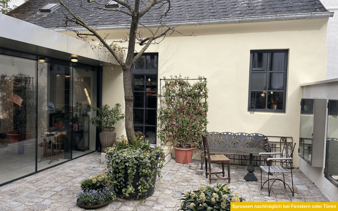 Ein idyllischer Innenhof mit antiken Metallmöbeln und vielen Pflanzen; die dunklen Fenster und Türen des hellen Hauses sind durch elegante Sprossen klassisch gegliedert.