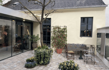 Ein gemütlicher Innenhof mit antiken Metallmöbeln und vielen Pflanzen; die Fenster des hellen Hauses sind durch nachträglich angebrachte dunkle Sprossen klassisch gegliedert.