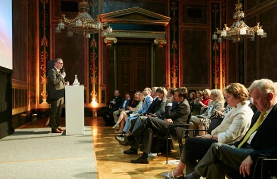 Offizielle Preisverleihung in einem historischen Salon mit roten Wänden, goldenen Stuckverzierungen und Kronleuchtern. Ein Redner hält eine Ansprache. Im Publikum aufmerksame Gäste in festlicher Geschäftskleidung.