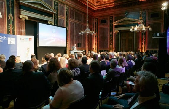 Festakt zur Staatspreis-KnewLEDGE-Verleihung in einem historischen, repräsentativen Saal. Ein Redner spricht am Stehpult, das Publikum sitzt in mehreren Reihen. Goldene Kronleuchter und rote Wände prägen das prunkvolle Ambiente.