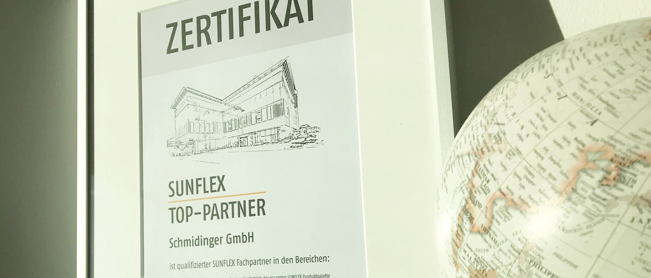 Zu sehen ist ein hell gerahmtes Zertifikat, das Fenster-Schmidinger als Sunflex Top-Partner auszeichnet. Neben dem Rahmen steht ein dekorativer Globus, beide Objekte werden von seitlichem Tageslicht angestrahlt.