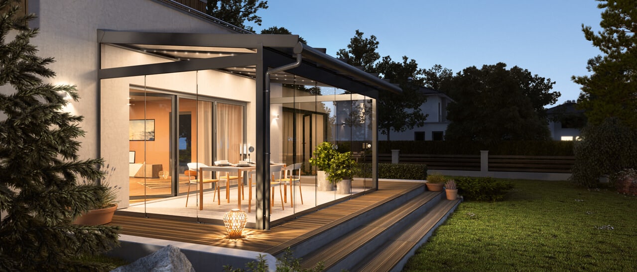 Zu sehen ist eine hochwertige moderne Terrassenüberdachung mit großzügigen Glasflächen, die den Außenbereich stilvoll und windgeschützt macht. Die Szene wirkt abends beleuchtet und gemütlich.