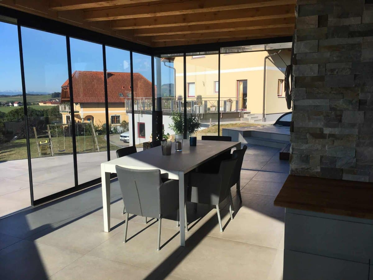 Überdachte Terrasse mit Essplatz und Glaselementen zum Schieben und Falten, dahinter Häuser und Landschaft