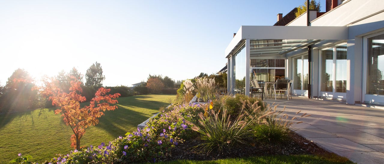 Großzügige Terrasse mit Glasüberdachung aus Aluminium, Gartenanlage mit bunten Blumen, herbstlicher Baum und Sonnenlicht im Hintergrund.