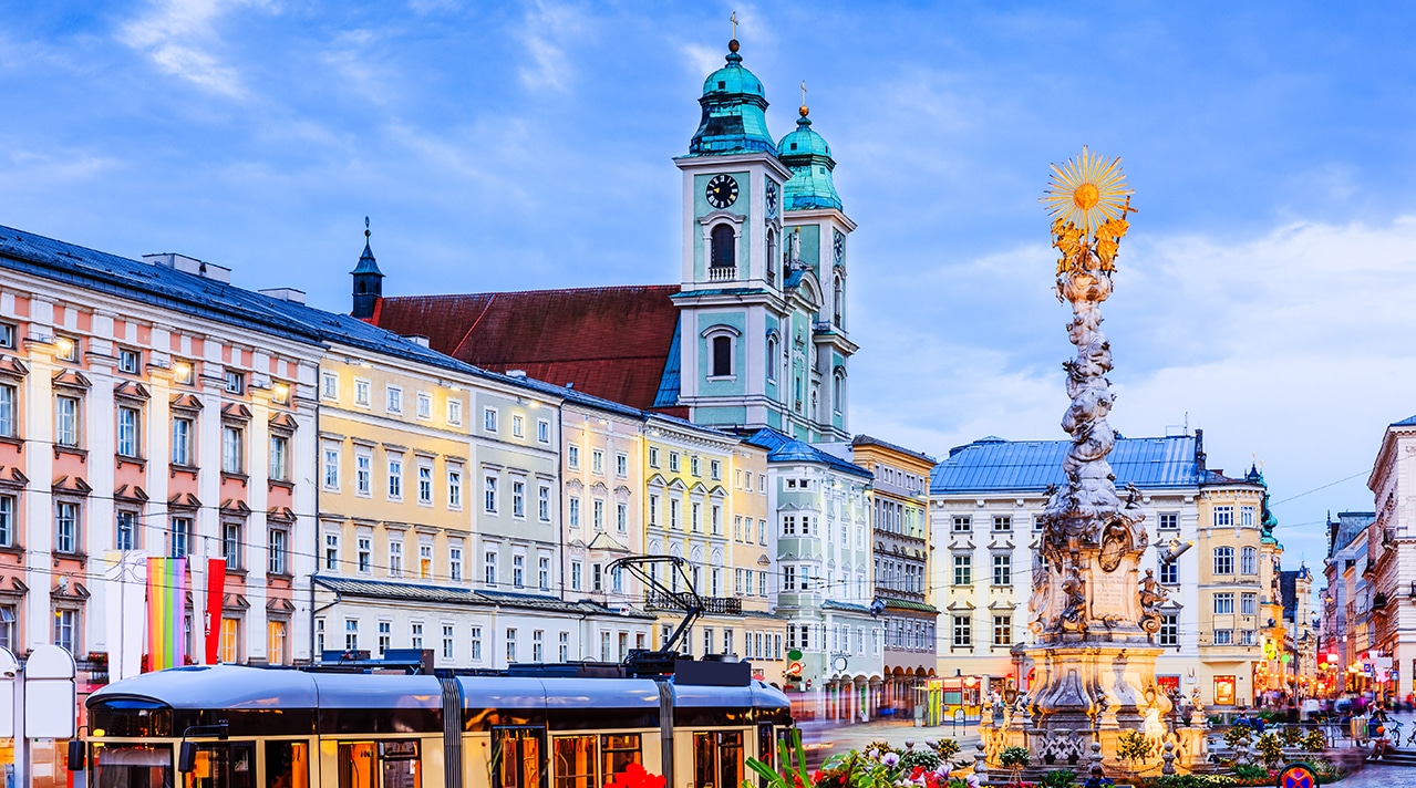 Historischer Hauptplatz mit bunten Fassaden, Straßenbahn, Blumenbeet und Dreifaltigkeitssäule – Symbol für Oberösterreichs Baukultur.