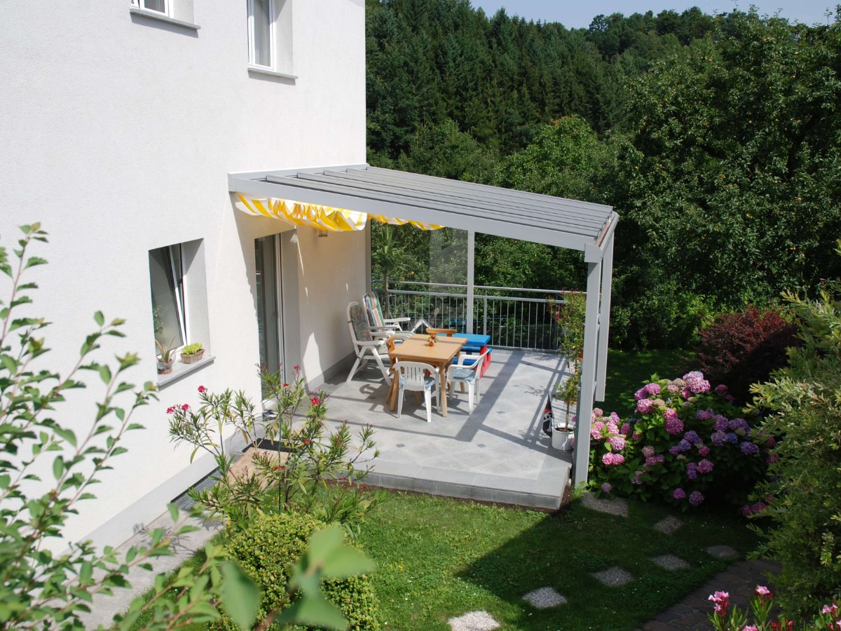 Terrassenüberdachung in RAL 9006 Weißaluminium mit Glasdach und gelber Markise über einer Terrasse im Grünen