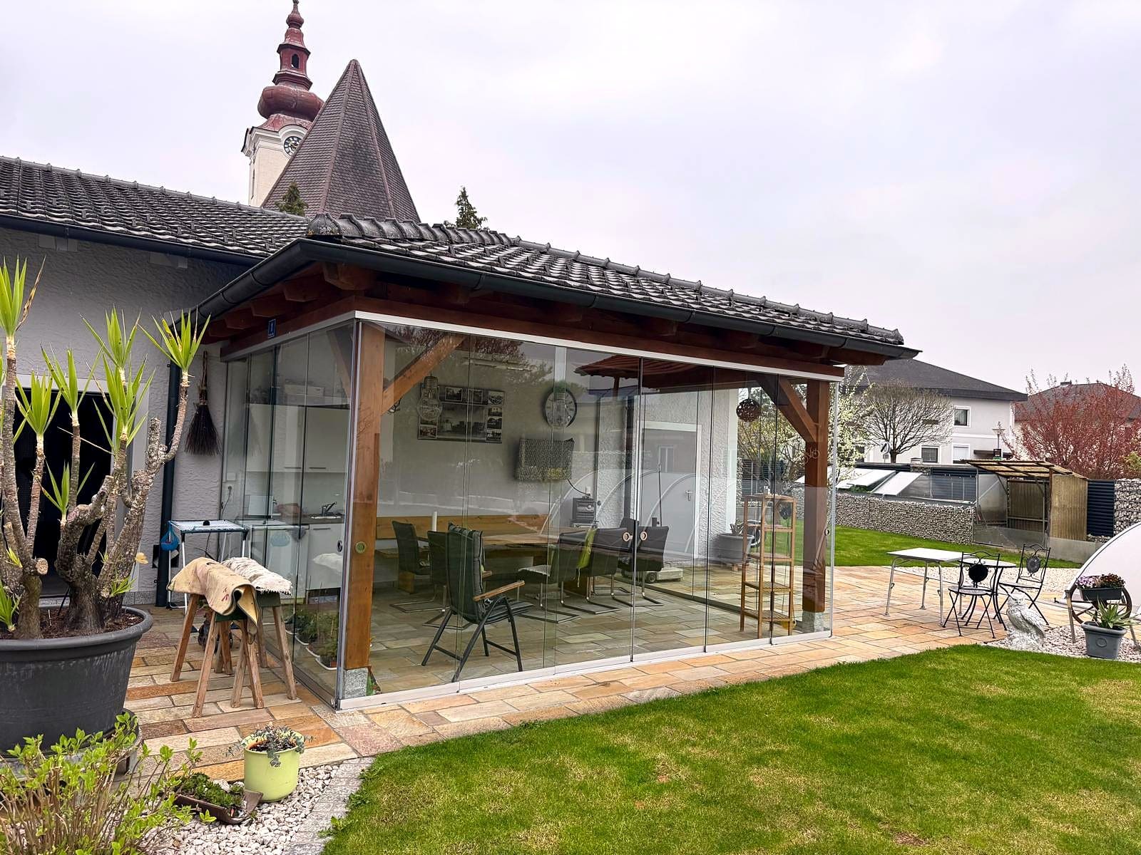 Terrassenverglasung mit rahmenlosen Ganzglas-Schiebetüren unter einem bestehenden Ziegeldach mit Holzstehern, Laufschienen in Alu natur eloxiert, Blick auf Esstisch und Garten, Oberösterreich