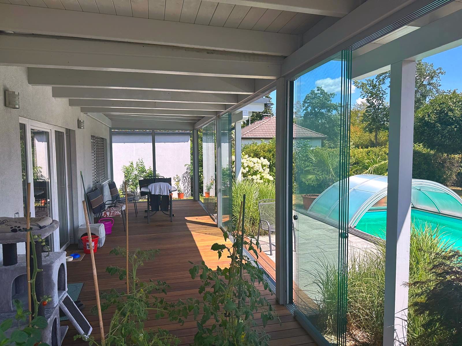 Terrassenverglasung mit rahmenlosen Ganzglas-Schiebetüren unter einem bestehenden Holzdach, Blick entlang der Holzterrasse zum Pool in Pasching