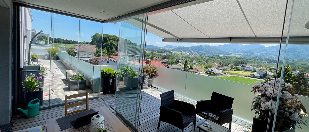 Überdachte Terrasse mit transparenten Glasschiebetüren. Sitzmöbel und Pflanzen stehen auf Holzboden, der Blick geht über eine grüne Hügellandschaft. Die Glaselemente lassen sich seitlich öffnen.