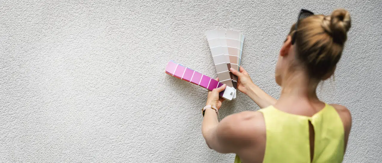 Junge Frau prüft RAL-Farben für Fenster und Türen an einer Hausfassade mit Farbpalette, um die beste Farbharmonie zu finden.