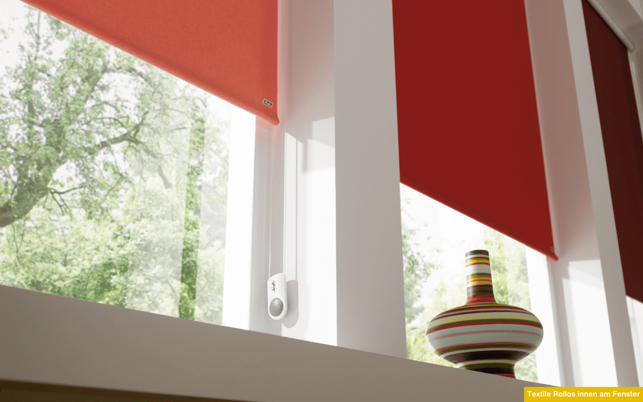 Das Foto zeigt rote, textile Innenrollos an modernen weißen Fenstern. Sie spenden Schatten, schützen vor Sonne und schaffen ein wohnliches Ambiente mit dekorativem Farbkontrast zu den hellen Fensterrahmen und Wänden.