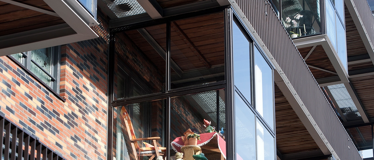 Das Bild zeigt einen verglasten Balkon mit Holzboden und gemütlicher Einrichtung. Durch die transparente Glasfassade bleibt der Bereich hell, geschützt und wirkt wie ein kleiner Wintergarten.