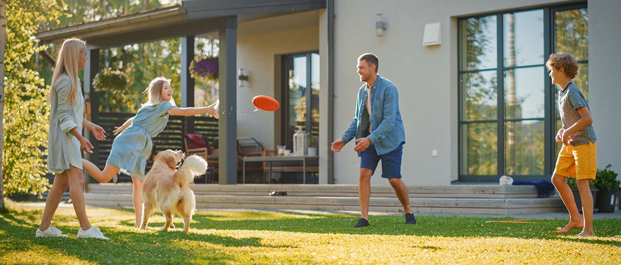 Eine Familie mit zwei Kindern und einem Hund spielt fröhlich im Garten vor einem modernen Haus mit großen grauen Fenstern. Sonnenlicht fällt warm auf die Szene.
