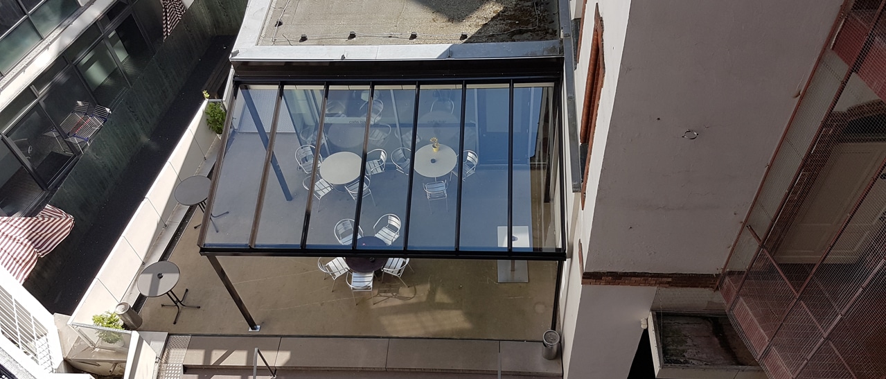 Wintergarten oder Terrassenüberdachung Vorteile Terrassenüberdachung Alu zeigt moderne Glasüberdachung mit Aluminiumrahmen über Sitzbereich im Innenhof.