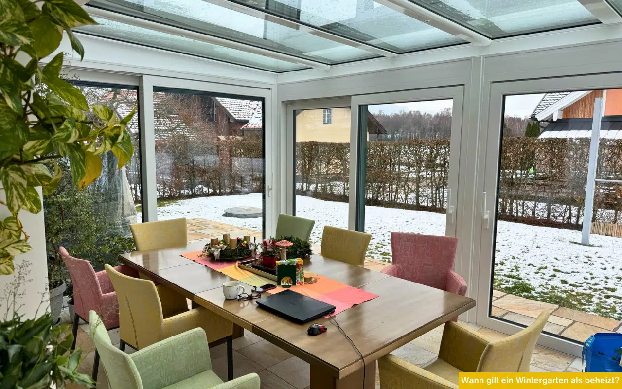 Wann gilt ein Wintergarten als beheizt zeigt verglasten Raum mit Tisch und Stühlen im Winter und Blick auf verschneiten Garten