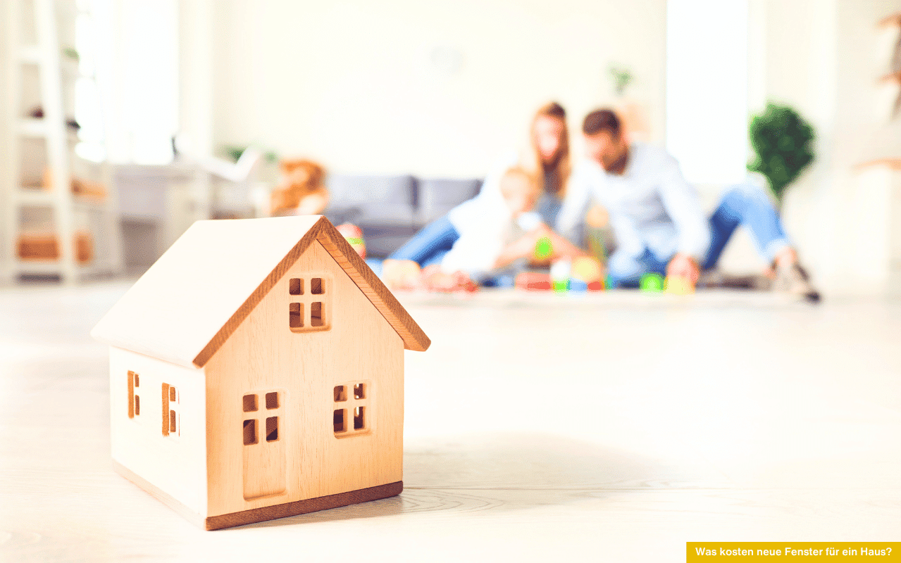 Was kosten neue Fenster für ein Haus? Symbolbild mit Holzmodell eines Hauses, im Hintergrund spielt eine Familie gemeinsam auf dem Boden.