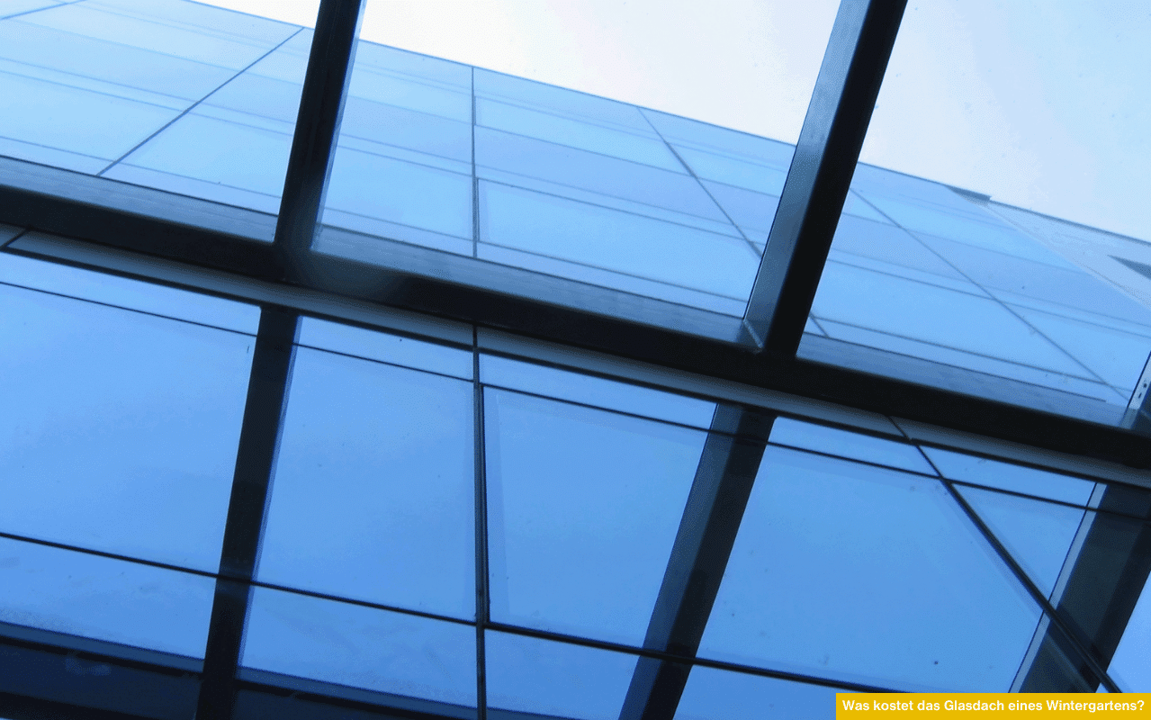 Teilansicht eines Glasdaches mit klaren, rechteckigen Glaselementen und stabilen Metallrahmen, fotografiert von unten gegen den Himmel.