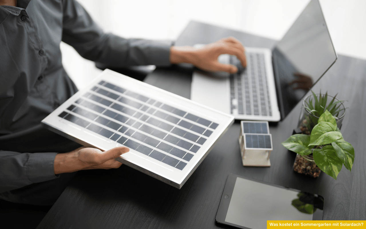 Eine Person sitzt am Schreibtisch, hält ein kleines Solarpanel in der Hand und arbeitet gleichzeitig am Laptop.