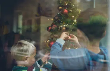 Kinder dekorieren Weihnachtsbaum hinter klarem Fensterglas sichtbar, festliche Stimmung.