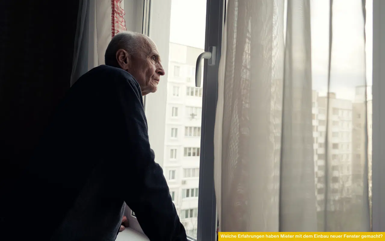 Ein älterer Mieter steht am neuen Kunststofffenster seiner Wohnung, schaut durch die halb geöffneten Vorhänge auf die Wohnsiedlung und wirkt dabei ruhig und nachdenklich – Symbolbild für persönliche Erfahrungen nach Fenstertausch.