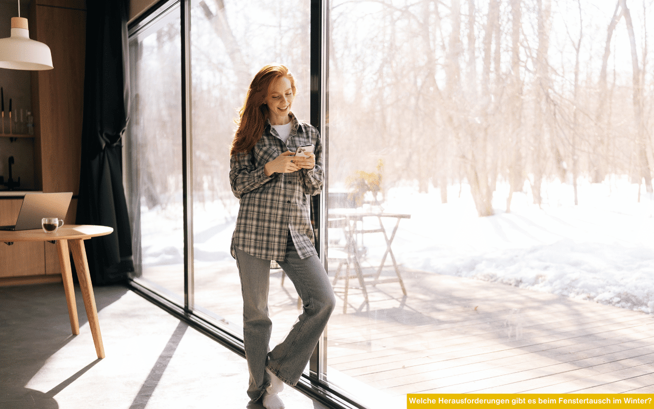 Das Bild zeigt eine Frau, die entspannt an einer großen Fenstertür steht und auf ihr Handy blickt. Draußen liegt Schnee, innen wirkt es hell und warm - Symbol für gute Isolierung moderner Fenster.